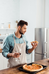 handsome young man in apron holding slice of homemade pizza and glass of wine