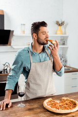 young man in apron eating homemade pizza in kitchen