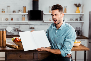 handsome young man holding blank cookbook and winking at camera in kitchen