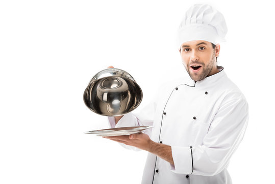 excited young chef taking of serving dome from plate and looking at camera isolated on white