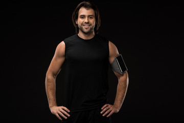 Fototapeta na wymiar athletic young man with armband and smartphone standing with hands on waist and smiling at camera isolated on black