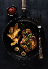 Roasted pork steak in frying pan with fried potatoes over dark background, top view.