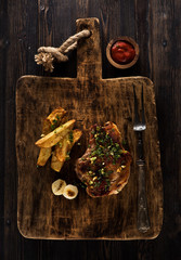 Roasted pork steak on a serving board with fried potatoes on a dark background, top view.