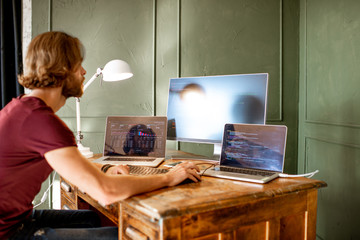 Young programmer writing a program code sitting at the workplace with three monitors in the office on the green wall background