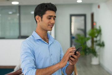 Content pensive young man reading Internet news on smartphone. Smiling Indian male manager searching for information using phone in office. Surfing net concept