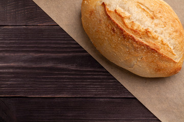 Simple minimalistic bread background. A white wheat loaf with a crispy fragrant crust lies on parchment over dark natural pine planks of the table. Parchment divides the frame diagonally.