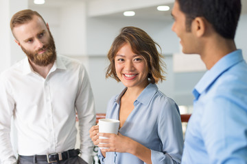 Multiethnic business group chatting during corporate meeting. Cheerful Asian office worker drinking coffee and smiling at camera while her colleagues talking. United team concept