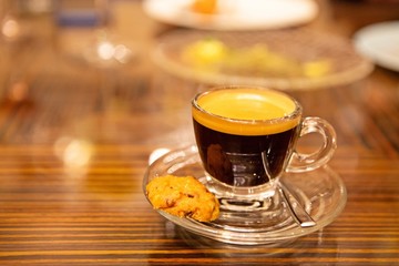 A single shot of freshly brew aromatic espresso coffee with crema in a clear glass cup with saucer and cookie. Good for caffeine boost in the morning. Selective focus. Close up. Warm light.