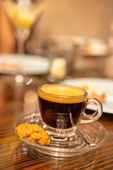 A single shot of freshly brew aromatic espresso coffee with crema in a clear glass cup with saucer and cookie. Good for caffeine boost in the morning. Selective focus. Close up. Warm light.