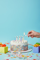 Partial view of woman lighting birthday cake on blue background with gifts and confetti