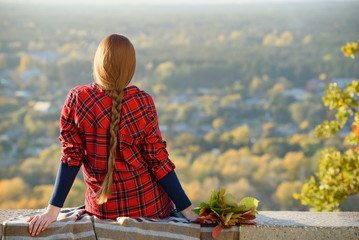 Young woman with long hair sits on a hill overlooking the city. Back view