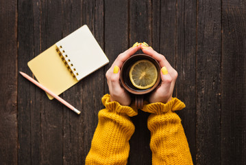 Mug of tea with lemon in a female hand, pencil and notepad. Wooden background. Top view