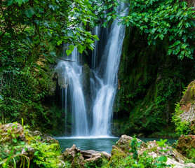 View to tall cascade falling to pond in green forest in long exposure