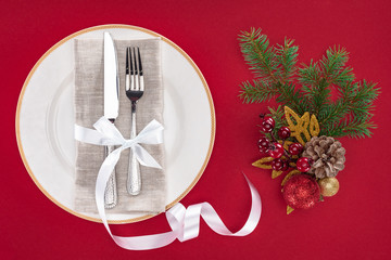 elevated view of fork and knife wrapped by ribbon on plate near christmas decorations isolated on red
