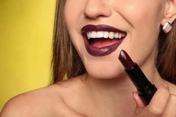 Beautiful young woman applying dark lipstick on color background, closeup