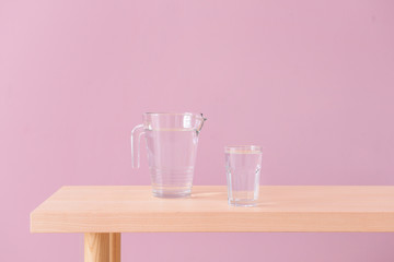 Glass and jug of fresh water on wooden table near color wall