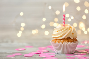 Delicious birthday cupcake with burning candle on grey table  against blurred lights