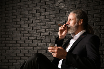 Senior man drinking whiskey and smoking cigar near dark brick wall
