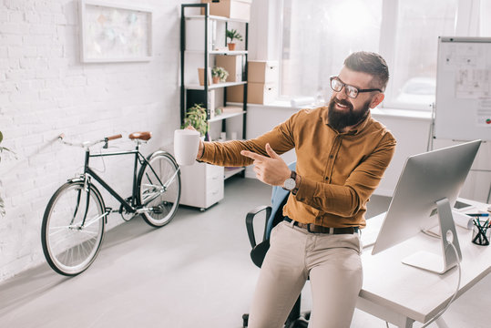 happy bearded adult businessman holding cup of coffee and pointing finger in exitement at workplace