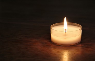 Fototapeta na wymiar Bright flame and white candle on dark background. Close up of a plain single tea candle or tea light with white wax on wooden table. Concept: light in darkness in times of festive, happy, sorrow, pray