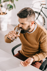 angle view of sick businessman sitting at office desk, holding medicine and drinking water at workplace