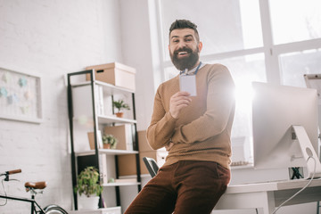 smiling bearded adult businessman sitting on office desk and holding blank security tag at workplace