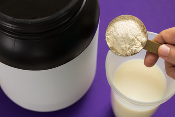 Whey Protein.  Vanilla flavour. Shaker with milk. Point of view, hand holding measuring scoop with vanilla flavour powder on violet background.