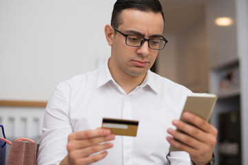 Concentrated young man using smartphone for online shopping. Portrait of mixed race man wearing glasses paying with credit card for online purchase. E-commerce concept