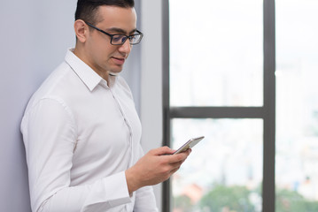 Portrait of smiling businessman using mobile phone at wall. Young Caucasian man wearing glasses reading message. Mobile communication concept