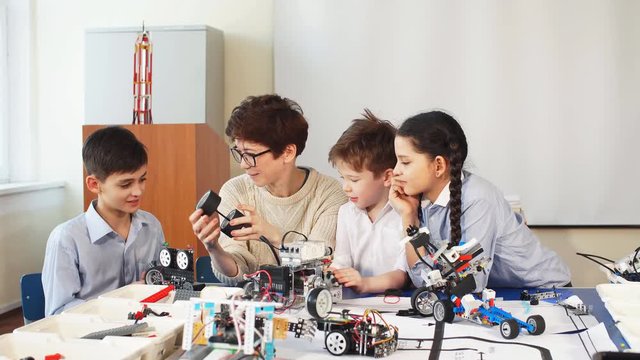 Group of happy kids with their female adult science prof with tablet pc computer programming electric toys and robots at robotics hobby club