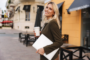 Plakat Image of cute woman 20s wearing jacket holding takeaway coffee and laptop, while walking through city street