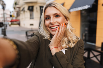 Image of beautiful woman smiling and taking selfie photo, while walking through city street