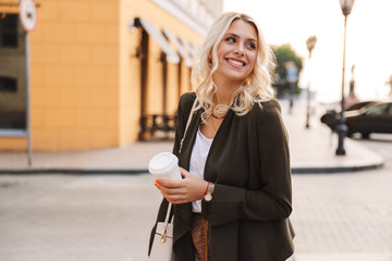 Image of beautiful woman wearing jacket holding takeaway coffee in paper cup, while walking through...