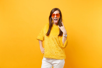 Funny crazy young woman in fur sweater and heart orange glasses screaming, showing rock-n-roll sign isolated on bright yellow background. People sincere emotions, lifestyle concept. Advertising area.