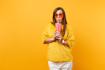 Shocked young woman in fur sweater and heart orange glasses drinking cola or soda from plastic cup isolated on bright yellow background. People sincere emotions, lifestyle concept. Advertising area.
