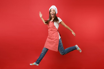 Housewife female chef cook or baker in striped apron white t-shirt, toque chefs hat isolated on red wall background. Full length portrait housekeeper woman jumping high up. Mock up copy space concept.