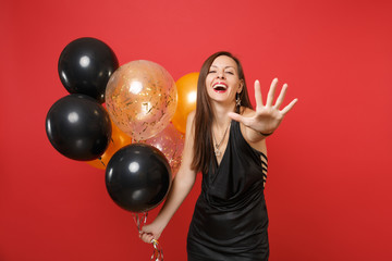 Fototapeta premium Laughing cheerful young woman in black dress pointing hand on camera and holding air balloons, celebrating isolated on red background. Women's Day Happy New Year birthday mockup holiday party concept.