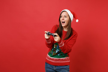 Amazed young Santa girl in Christmas hat keeping mouth wide open playing video game with joystick isolated on red background. Happy New Year 2019 celebration holiday party concept. Mock up copy space.