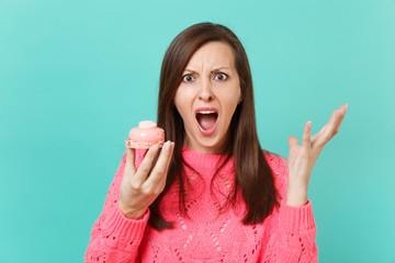 Shocked angry young woman in knitted pink sweater spreading hands screaming hold cake isolated on blue wall background, studio portrait. People sincere emotions, lifestyle concept. Mock up copy space.
