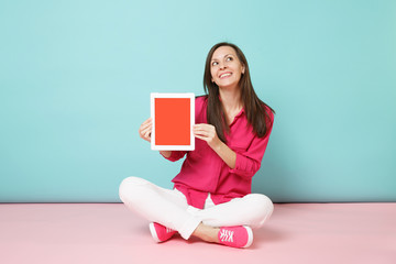 Full length portrait woman in rose shirt blouse, white pants sitting on floor hold tablet pc...