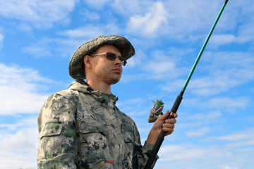 The suntanned caucasian man is wearing a camouflage suit holding the fishing spinning with reel in his hand against a background of the blue sky. The pensive fisherman is looking into a distance.