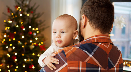 family, childhood, holidays and people concept - close up of father holding baby son at home over christmas tree lights background