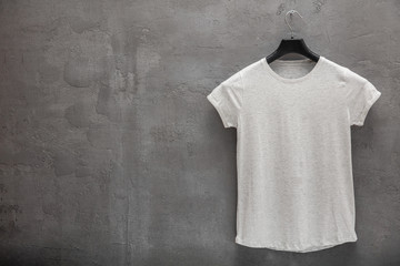 Front side of female grey melange cotton t-shirt on a hanger and a concrete wall in the background....