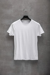 Front side of male white cotton t-shirt on a hanger and a concrete wall in the background. T-shirt...