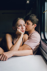 Mood portrait of young lifestyle couple of lovers traveling in train. Beautiful loving pair hugging, kissing, telling secret near window. Boy and girl in love.  Friends in journey on vacation.