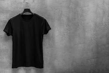 Front side of male black cotton t-shirt on a hanger and a concrete wall in the background. T-shirt without print and copyspace for your text on right side