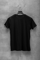Back side of male black cotton t-shirt on a hanger and a concrete wall in the background. T-shirt...