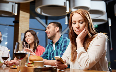 technology, lifestyle and people concept - bored woman dining with friends and messaging on smartphone at restaurant