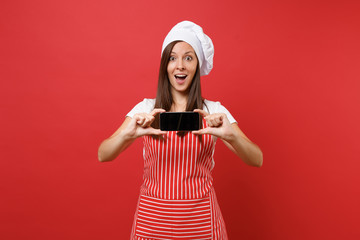 Housewife female chef cook or baker in striped apron white t-shirt toque chefs hat isolated on red wall background. Woman hold cellphone blank screen for promotional content Mock up copy space concept