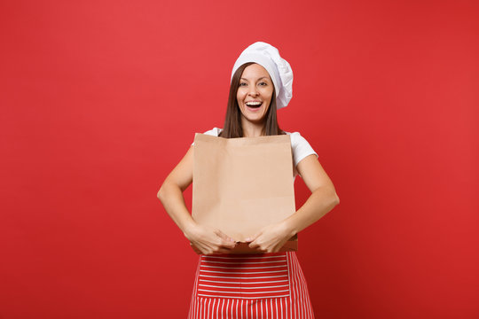 Housewife female chef cook baker in striped apron t-shirt toque chefs hat isolated on red wall background Woman hold in hand brown clear empty blank craft paper bag takeaway Mock up copy space concept